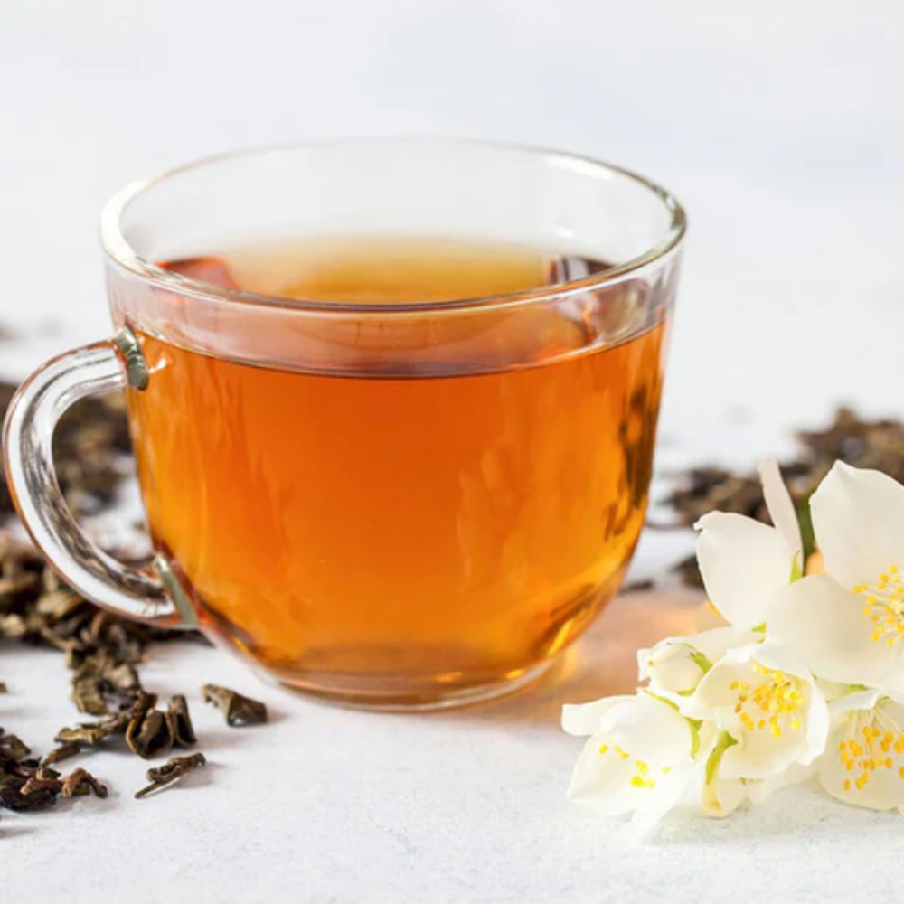 Clear glass cup filled with tea, surrounded by dried tea leaves and white flowers on a light background
