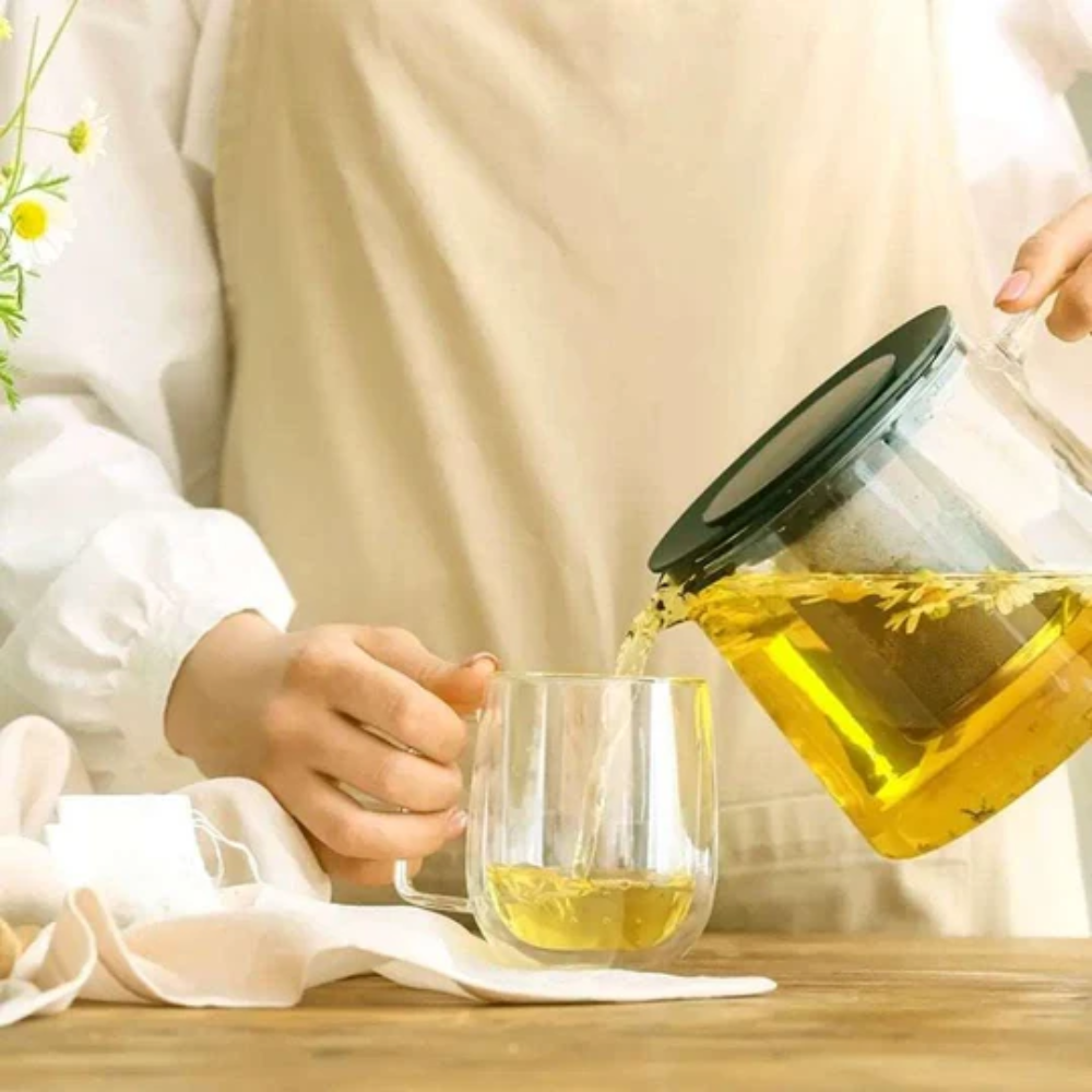 Person pouring tea from a glass teapot into a clear mug on a wooden table.