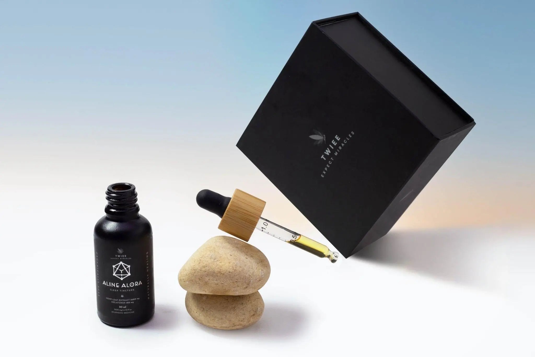 Twiee Aline Alora tincture bottle with dropper above two beige stones and a black box.